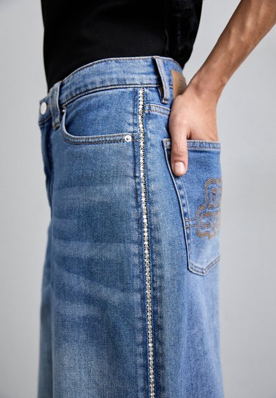 Mėlyna džinsinė sijonas su šonine kišene, sidabro detalės išsiuvinėjimo linijoje ir siuvinėjimu ant galinės kišenės.