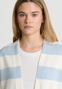 Dryžuotas švarkas šviesiai mėlynos ir grietinėlės spalvų, pasižymintis minkšta mezgimo tekstūra ir atidarytu priekio dizainu, dėvimas virš raibalių baltų marškinėlių.