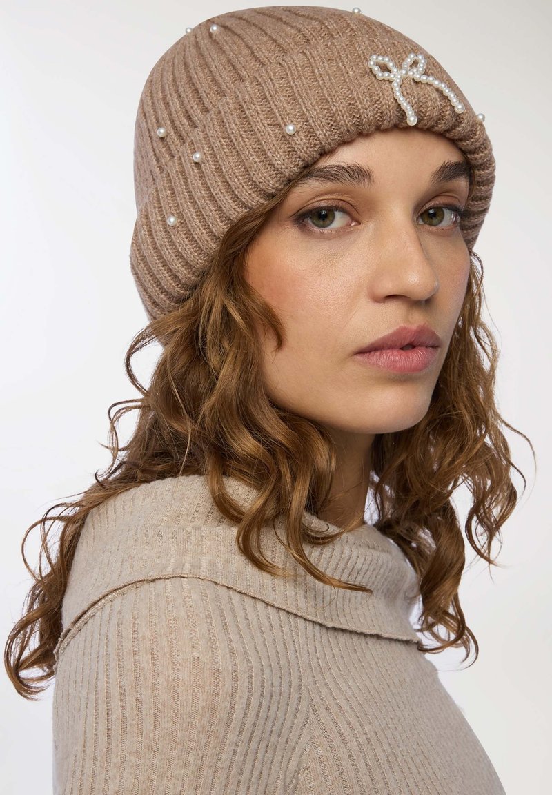 Beanie beige lavorato a maglia con texture a coste, decorato con piccole perle e un fiocco impreziosito da perline sul davanti.