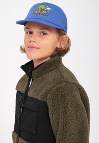 Casquette en maille bleue avec un design graphique, associée à une veste en polaire marron avec un panneau avant noir et une poche zippée. Tissu texturé visible.