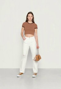 Jovem mulher vestindo uma t-shirt cropped às riscas castanhas, jeans brancos rasgados, ténis castanhos, segurando uma pequena bolsa castanha com alças de corrente.
