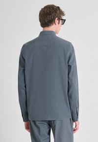 Camisa de manga larga en tejido gris medio, textura suave, cuello clásico, puños abotonados, sutil marca en el cuello, llevada con gafas de sol.