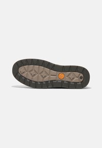 Gummisohle mit einem strukturierten Design, das tiefe Profilrillen aufweist, in einem beige und schwarzen Farbschema, und einem runden orangen Logo in der Mitte.