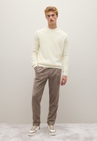 Cremfarbener Rippstrickpullover, hellbraune maßgeschneiderte Hose und weiße Sneakers mit beigen Akzenten. Das Model steht vor einem einfarbigen Hintergrund.