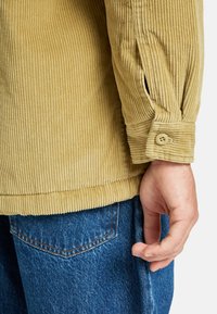 Camicia di velluto a coste beige con ampie costripre, dettagli con polsini abbottonati, abbinata a jeans in denim blu con una tasca posteriore visibile.