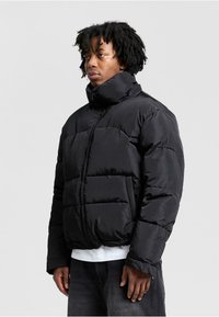 Sort puffet jakke med høj krave, ziplukning og overdimensionerede quiltede sektioner. Teksturstof og synlige frontlommer.