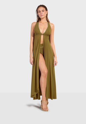 Žena u maslinasto zelenoj halter maxi haljini s prednjim prorezom koji otkriva odgovarajuće bikini gaćice i zlatne sandale, stoji na sivoj pozadini.