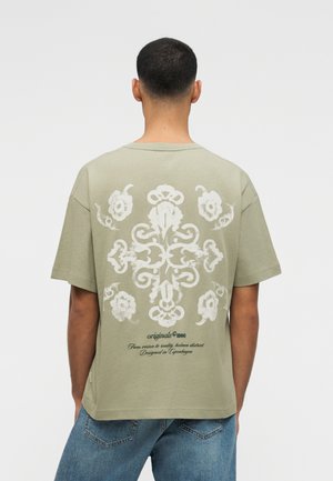 Άνδρας με κοντά μαλλιά φοράει χακί μπλουζάκι με λευκό περίτεχνο λουλουδάτο σχέδιο και κείμενο στην πλάτη, συνδυασμένο με μπλε τζιν.