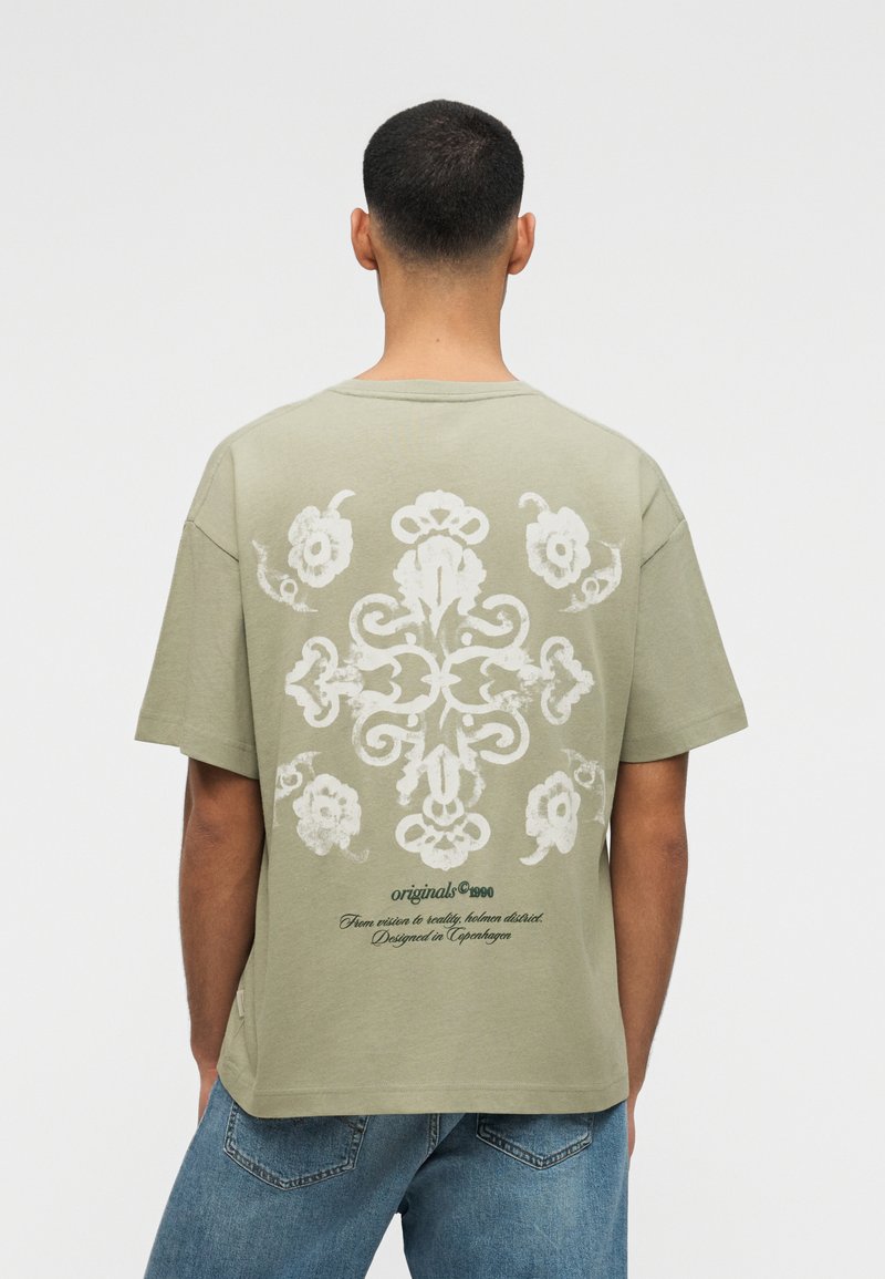 Mężczyzna o krótkich włosach w khaki koszulce z białym ozdobnym wzorem kwiatowym i napisem na plecach, w połączeniu z niebieskimi dżinsami.