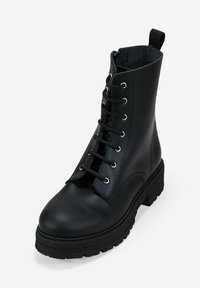 Bota de combate negra de cuero con cordones, suela de goma gruesa y tirador en el tobillo, fotografiada sobre un fondo blanco liso.