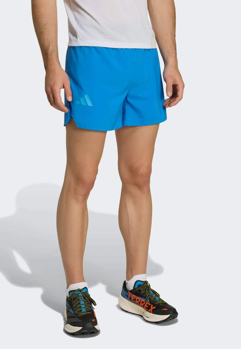 Homme portant un short de sport bleu vif, des chaussettes blanches à la cheville et des chaussures de course noires et bleues avec un marquage orange "TERREX".