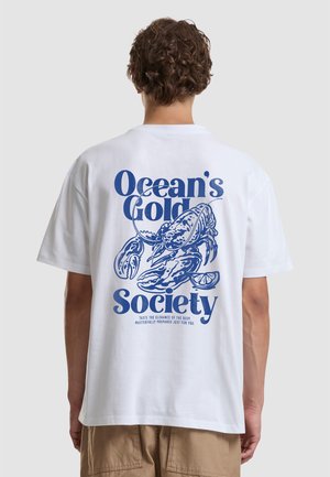 Személy fehér pólót visel, amelyen kék homár grafika és az „Ocean's Gold Society” felirat található a hátoldalon, sima háttér előtt áll.