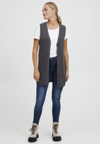Grijze gebreide vest met een textuurpatroon, met vijf knopen, gedragen over een wit T-shirt en gecombineerd met blauwe jeans en beige laarzen.
