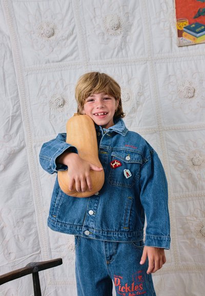 Jeune garçon en veste et pantalon en jean souriant, tenant une grosse courge butternut devant un fond blanc matelassé.