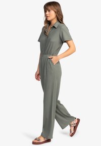 Olijfgroen jumpsuit met korte mouwen, een knoopsluiting aan de voorkant, elastische taille en zijzakken; gecombineerd met sandalen met gekleurde banden.