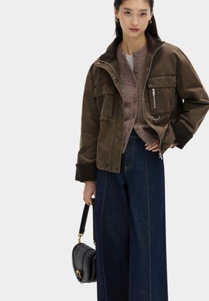 Frau trägt eine braune, geknöpfte Strickjacke unter einer olivgrünen Zip-up-Jacke, weit geschnittene dunkelblaue Jeans und hält eine schwarze Handtasche mit goldener Hardware.