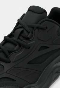 Μαύρο αθλητικό παπούτσι με υφασμάτινο επάνω μέρος που συνδυάζει δίχτυ και συνθετικά επικαλύμματα, στρογγυλεμένη μύτη και επίπεδους κορδόνια. Διακριτικές σχεδιαστικές λεπτομέρειες σε όλο το παπούτσι.