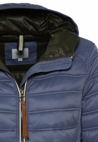 Veste matelassée bleue avec doublure intérieure noire, capuche et fermeture éclair argentée avec un curseur en cuir marron.