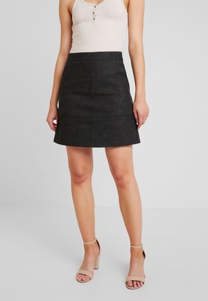 Jupe en simili cuir noir en forme A avec une texture lisse, une taille ajustée et des coutures visibles, associée à des sandales à talons beige.