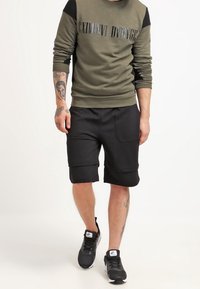 Olijfgroen sweatshirt met zwarte schouderaccenten en metallic tekst, gecombineerd met zwarte shorts en zwarte sportschoenen.