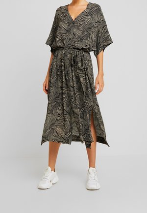 Vrouw draagt een midi-jurk met een abstract zwart-wit patroon, een V-hals, brede mouwen, ceintuur in de taille, zijsplit en witte sneakers.