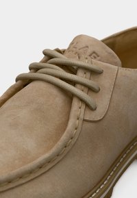 Κοντινό πλάνο μπεζ παπουτσιού από καστόρι με στρογγυλεμένη μύτη, χοντρές μπεζ κορδόνια, λεπτομερείς ραφές και μερικό ανάγλυφο λογότυπο στη γλώσσα.