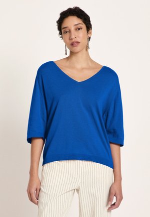 Frau mit kurzen lockigen Haaren, die ein blaues V-Ausschnitt-Oberteil und weiße, fein gestreifte Hosen vor schlichtem Hintergrund trägt.