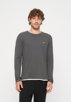 Homme aux cheveux bouclés portant un t-shirt gris foncé à manches longues et un pantalon noir, debout devant un fond clair uni.