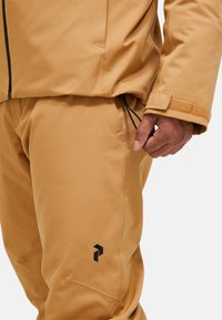 Completo outerwear color marrone chiaro con una texture liscia. La giacca ha una chiusura con zip e un orlo regolabile; i pantaloni hanno tasche laterali e un dettaglio con logo.