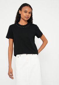 Donna con capelli ricci che indossa una maglietta nera con borchie e scollo rotondo e una gonna bianca a vita alta, posando con una mano sui fianchi davanti a uno sfondo neutro.