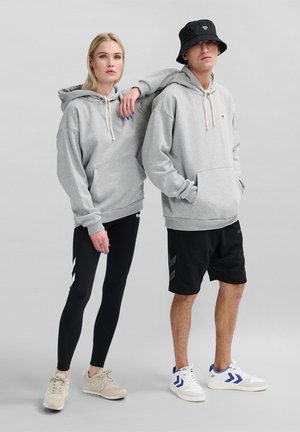 Übergroße graue Kapuzenpullover mit Kordelzug, kombiniert mit schwarzen Leggings mit weißen Zickzack-Mustern und schwarzen Shorts mit grauen Akzenten. Sneakers in Beige und Weiß.