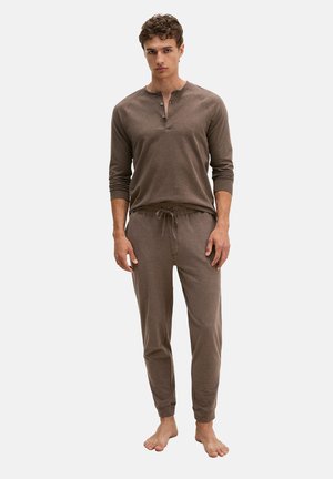 Jonge man staat op blote voeten, draagt een bruine henley met lange mouwen en bijpassende joggingbroek met trekkoord tegen een effen achtergrond.