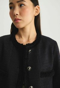 Veste noire texturée avec un col rond à franges, des boutons en argent et des poches avant. Le tissu semble tissé et consistant.
