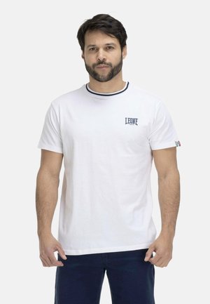 Homme aux cheveux foncés et à la barbe portant un T-shirt blanc à manches courtes avec le logo « LEONE 1947 » sur la poitrine et un pantalon foncé.