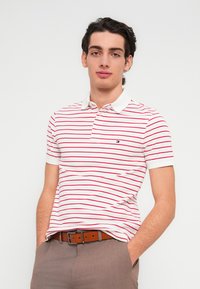 Λευκό πόλο μπλουζάκι με κόκκινες ρίγες, κοντά μανίκια και γιακά. Διαθέτει μικρό λογότυπο στο αριστερό στήθος. Καφέ ζώνη και ανοιχτό καφέ παντελόνια.