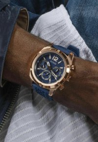 Orologio con cinturino in gomma blu, cassa in oro rosa, quadrante textured blu navy, dettagli in argento e grande numero 12, indossato su un polso.