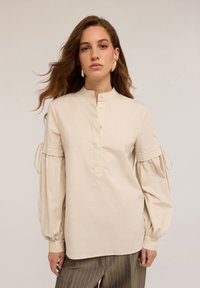 Beige, langærmet bluse med mandarinkrave, knaplukning og pufferede ærmer med snore. Glat stof med en let struktur.
