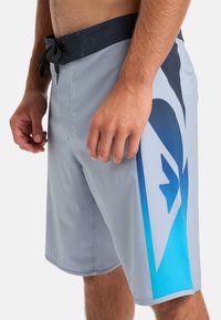 Svetlošedé plavky s tmavým pásom, s modro-tyrkysovým gradientným dizajnom na boku. Materiál sa zdá byť hladký a rýchloschnúci.