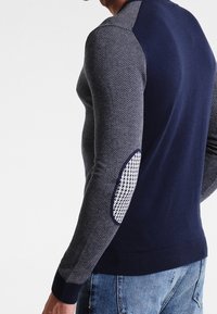 Mörkblå tröja med texturerade ärmar i fiskbensmönster, ribbade muddarna och ett distinkt vitt accent på armbågen.