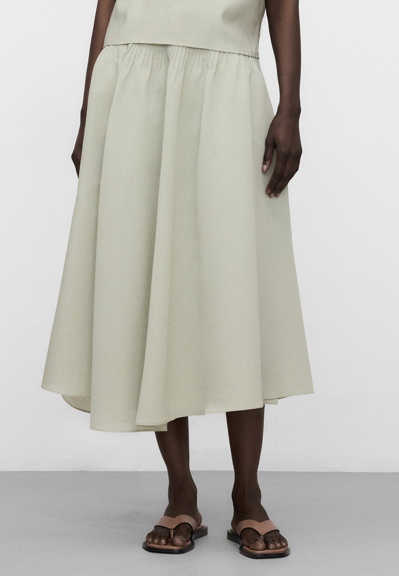 Mannequin portant une jupe midi beige clair froncée à la taille, associée à des sandales plates marron clair, posant devant un fond uni.