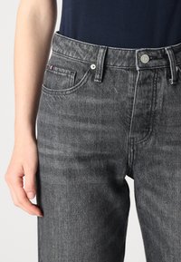 Jean en denim gris avec une texture légèrement décolorée, design à cinq poches et un petit patch logo coloré sur la taille.
