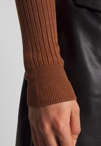 Brązowy sweter z prążkowanym dzianinowym rękawem z dopasowanymi mankietami, zestawiony z gładkim czarnym skórzanym tłem, podkreślający kontrast faktur.