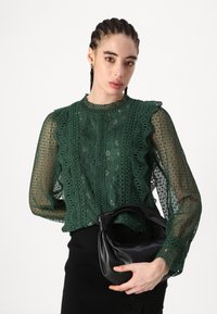 Blusa verde in pizzo con motivi intricati, bordi arricciati e maniche lunghe trasparenti; abbinata a una borsa nera morbida con manico a nodo.