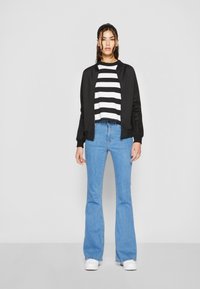 Zwarte bomberjack over een wit en zwart gestreept crop top, gecombineerd met lichtblauwe flare jeans en witte sneakers. Soepele texturen en casual ontwerp.