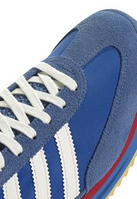 Sininen urheilukenkä, jossa mokka- ja tekstiilimateriaaleja. Sisältää valkoisen kolmiraidallisen kuvion, teksturoidun pinnan ja punaisen korostuksen pohjassa.