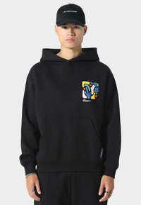 Sudadera con capucha negra que presenta un bolsillo frontal, puños acanalados y un diseño abstracto colorido en el lado izquierdo del pecho. Textura de tela suave.
