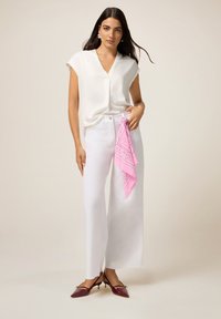 Femme portant une blouse blanche et un pantalon large blanc, avec un bandana rose suspendu à la poche, associé à des talons aiguilles rouge foncé.