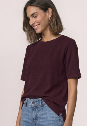Kvinde smilende, iført en tekstureret mørkelilla kortærmet top stoppet ned i lyseblå jeans med guldringeøreringe.