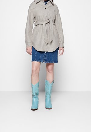 Frau trägt beigen Mantel mit Gürtel, blauen Jeansrock und hellblaue Cowboy-Stiefel, steht vor weißem Hintergrund.