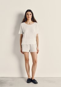 Ženska stoji ob bež steni, oblečena v široko belo majico, svetlo sive kratke hlače in črne čevlje brez vezalk, gleda naravnost predse.